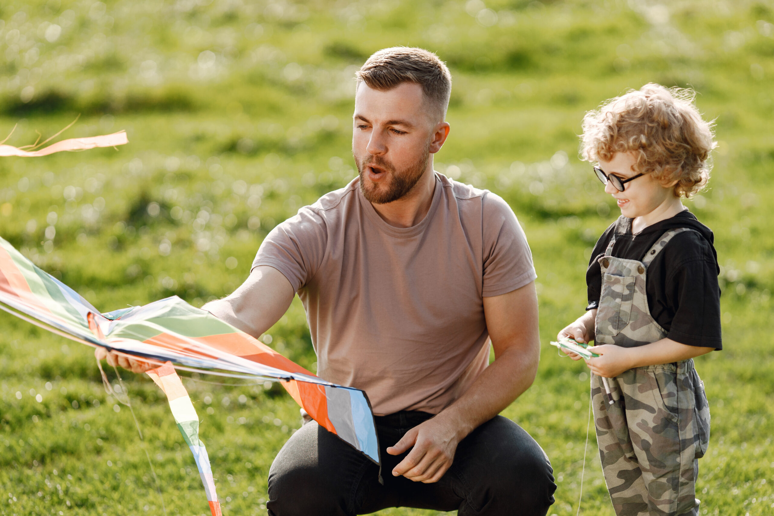 Single caucasian dad and child boy play and laugh together. Father and son playing with a kite and having fun on summer park outdoor. Curly toddler boy has a glasses.