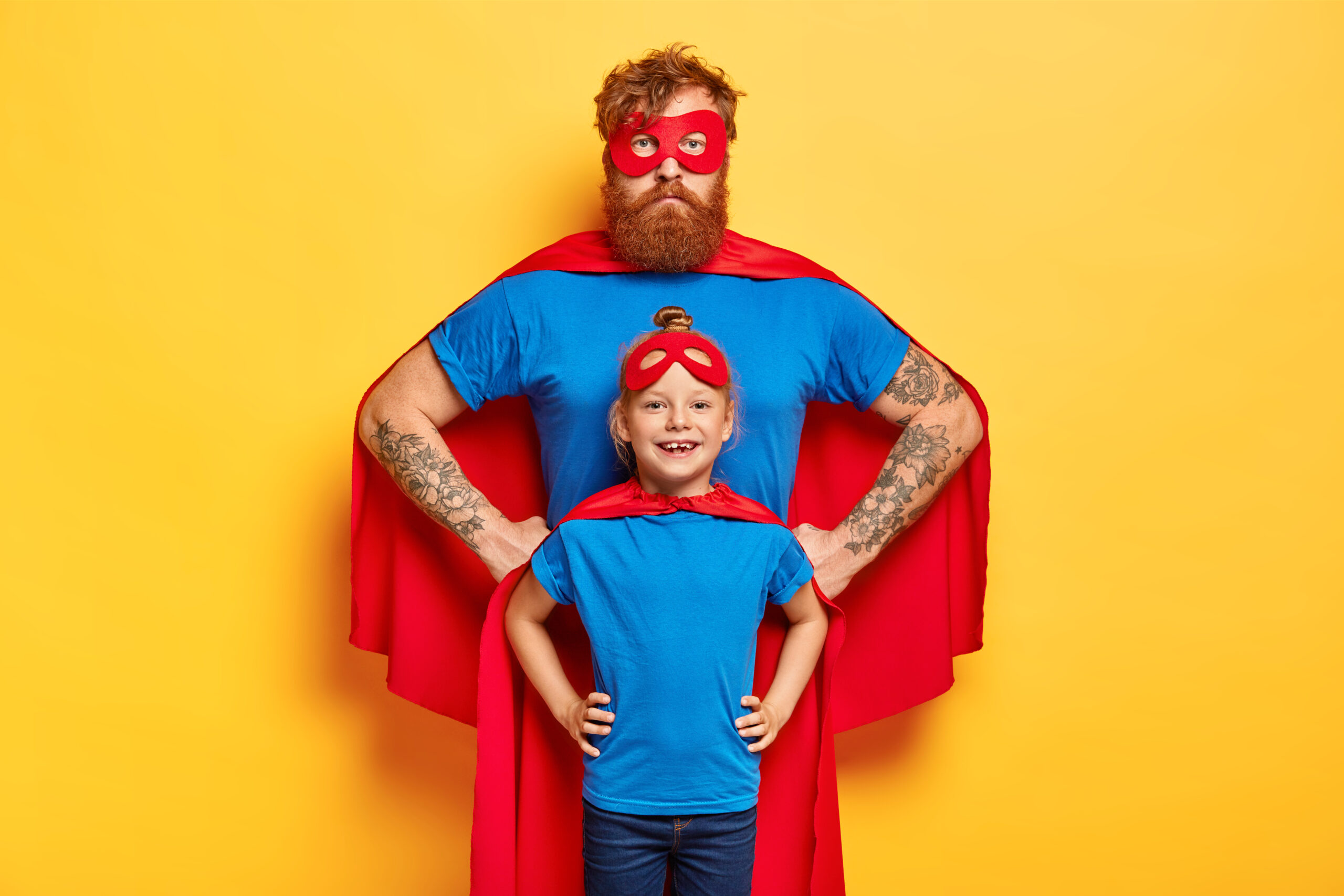 Photo of confident ginger father and daughter keep hands on waists, pretend being superhero and defend people, wear special costumes, isolated on yellow background. Family team ready to work