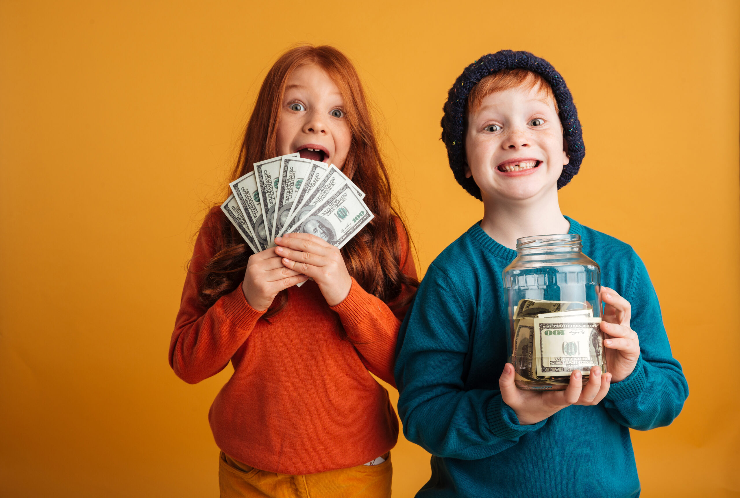 Photo of two excited little redhead children with freckles standing isolated over yellow background wearing warm hats. Looking camera holding money.