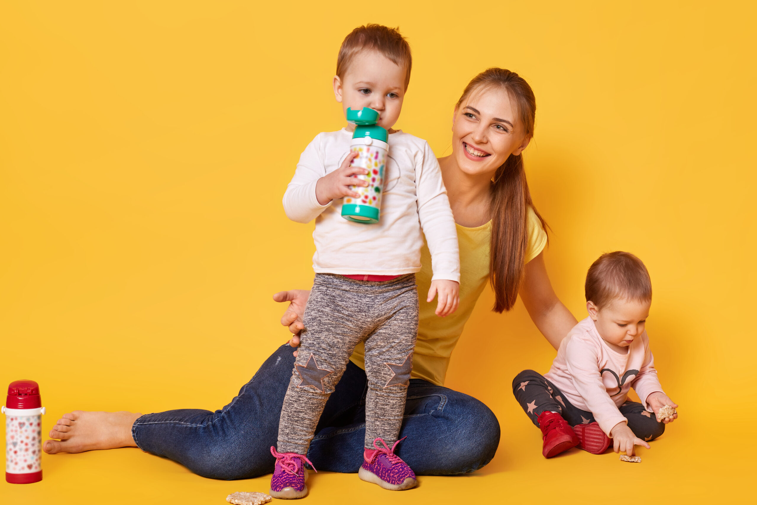 Image of loving attractive mother looks after her little children, twins playing with mommy. Playful kids drink tasty baverage from her bootle while her sister eating coockies. Infants are hungry.