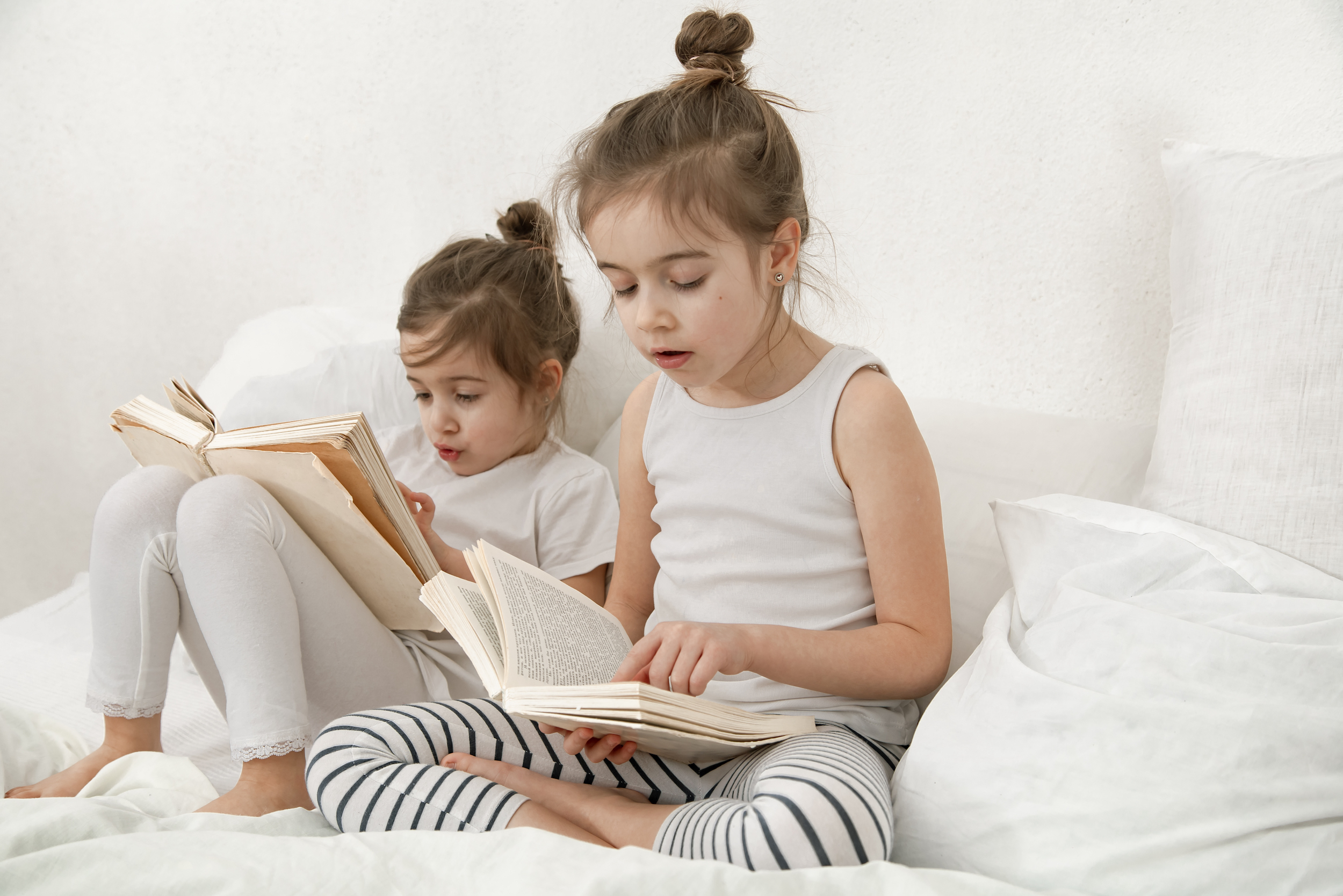 Two cute little sister girls are reading a book on the bed in the bedroom. The concept of family values and children's friendship.