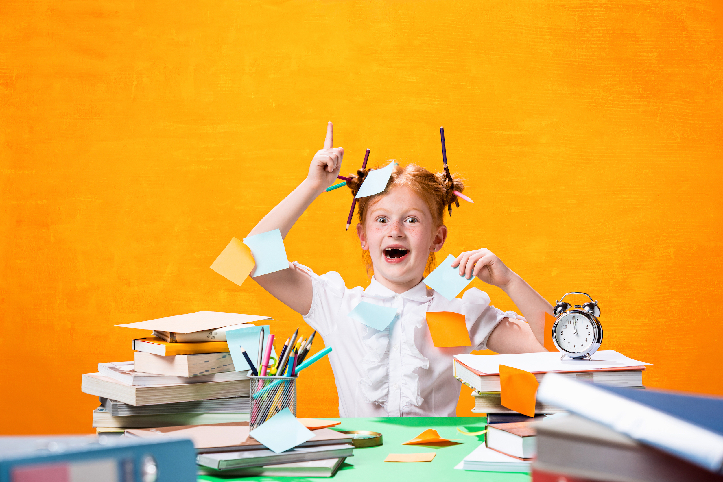 The Redhead teen girl with lot of books sitting at table on orange studio background. The education and back to school concept