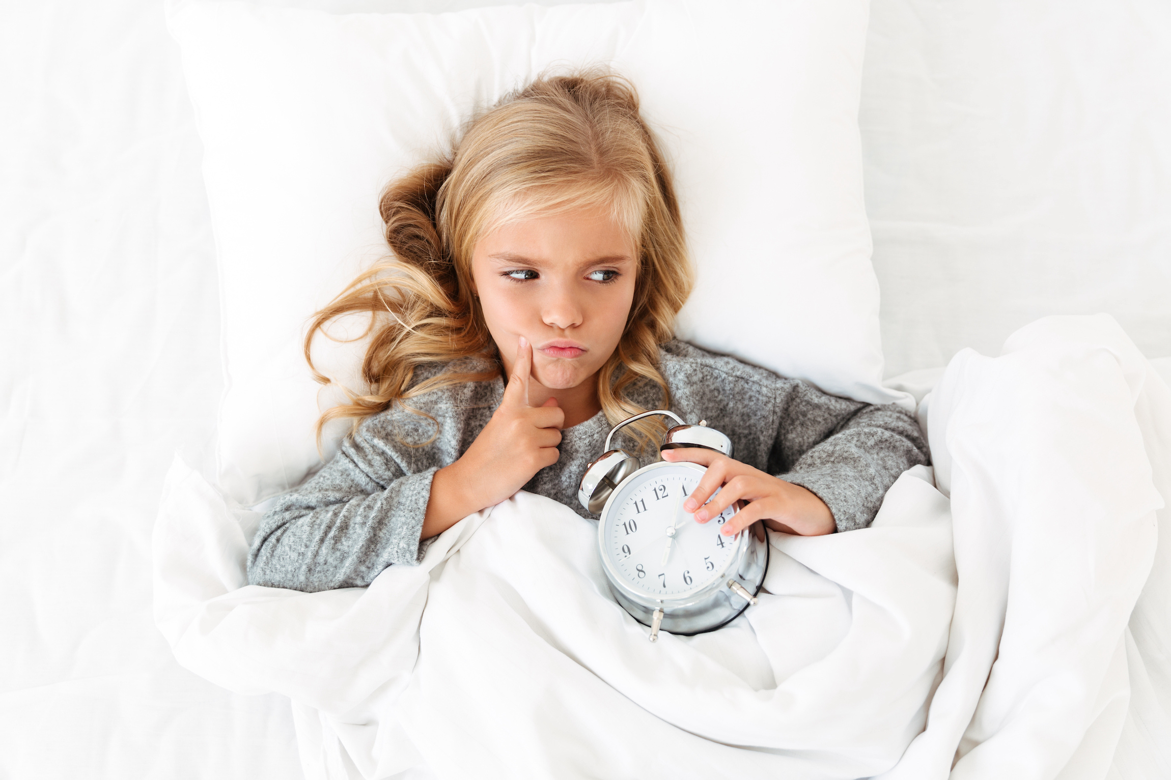 Top view of thoughtful little girl lying in bed with alarm clock, looking aside